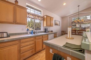 A kitchen with wooden cabinets, a window with a view, a coffee maker, and a dining area with chairs. - Private Hot Tub + Fireplace, 5 Min to Slopes, Fenced Yard, Sleeps 8 (Big Bear)