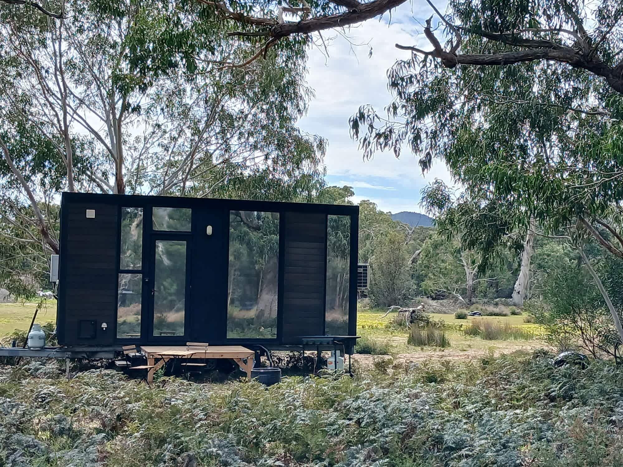 Une petite cabane aux couleurs sombres, dotée de grandes fenêtres et entourée d’arbres et de végétation.