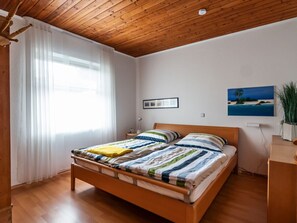 A bedroom with a wooden ceiling, a bed with striped bedding, a window with white curtains, and a painting on the wall. - Holiday Apartment With 1 Bedroom (Wunstorf)