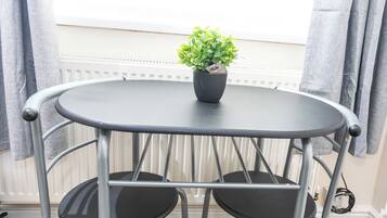 Una mesa metálica redonda con dos sillas, una planta en maceta y una ventana con cortinas.