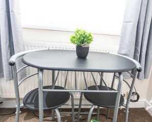Una mesa metálica redonda con dos sillas, una planta en maceta y una ventana con cortinas.