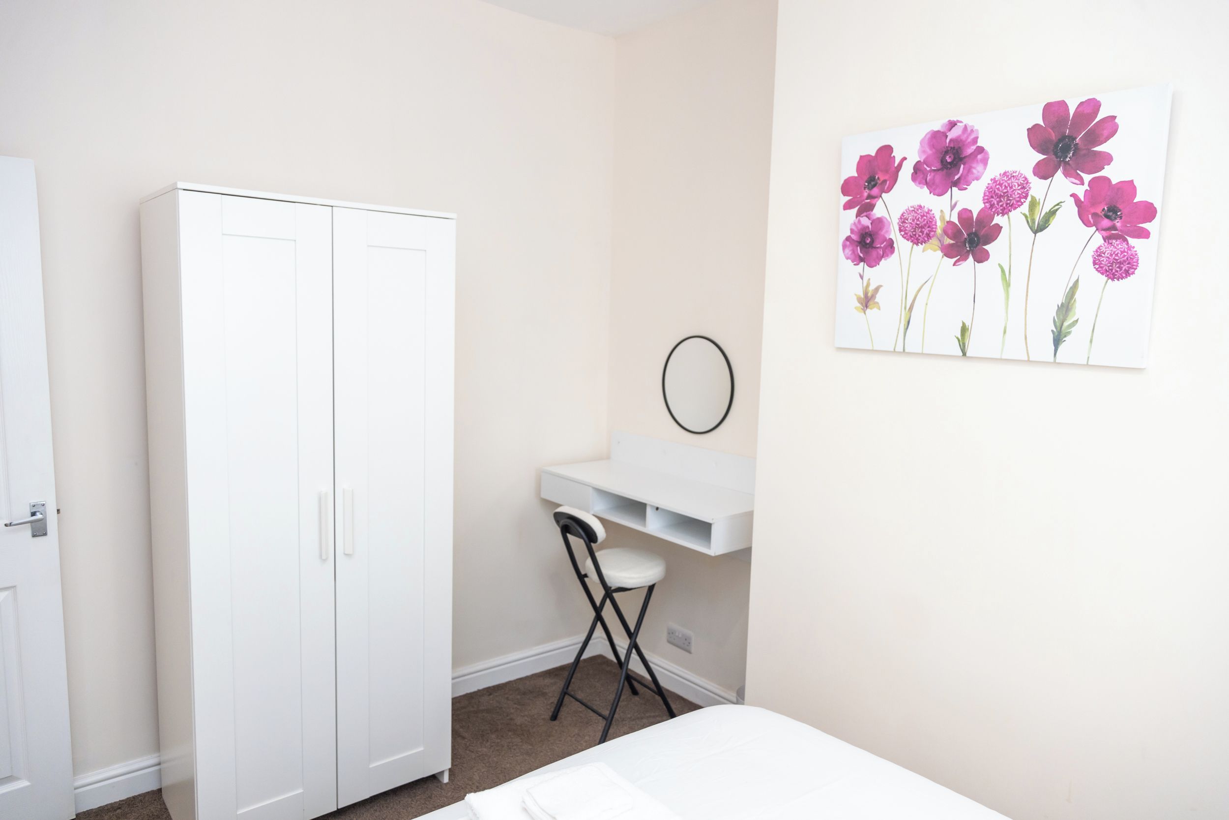 A bedroom with a white wardrobe, a desk with a chair, and a wall-mounted mirror.