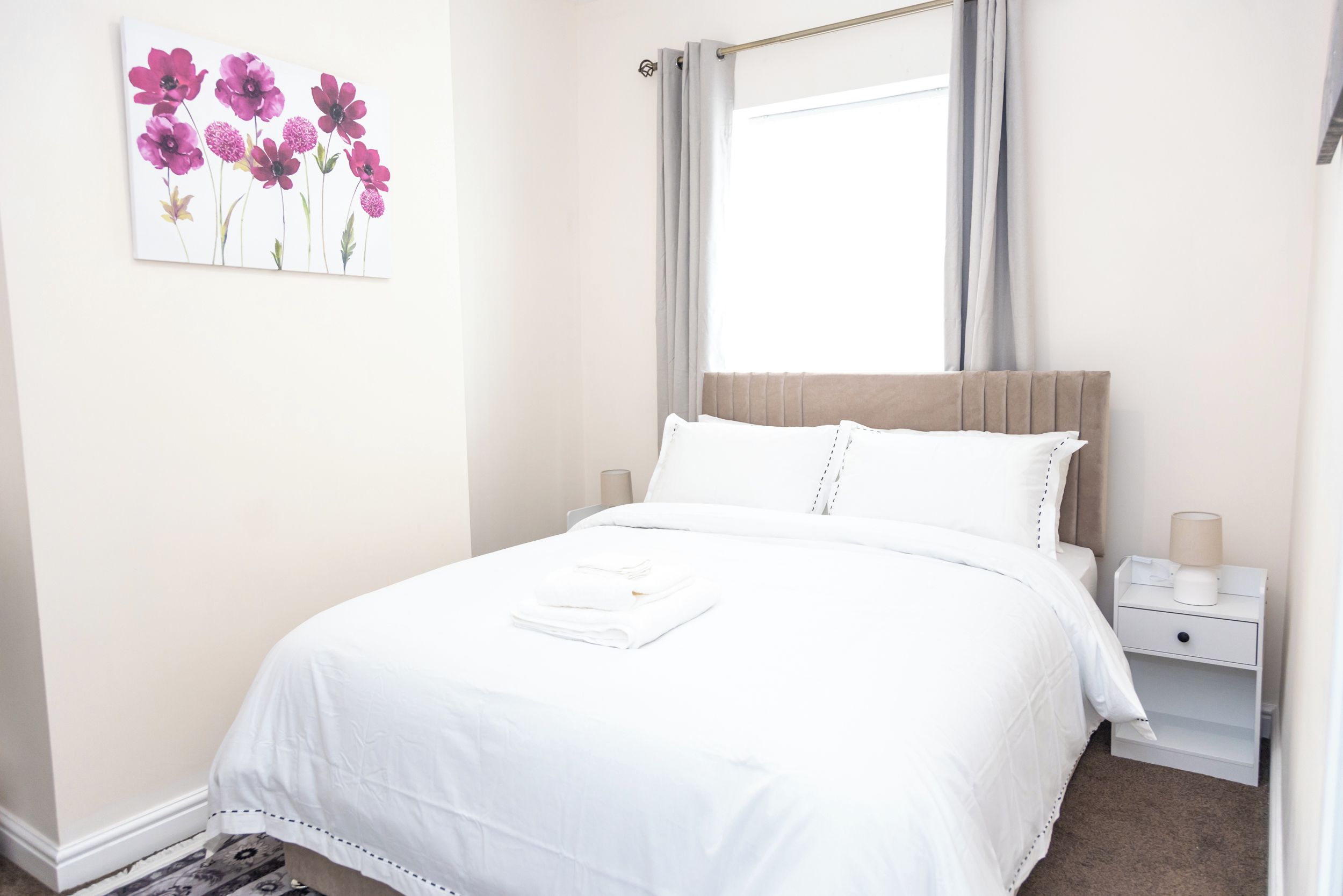 A neatly made bed with white linens, a bedside table with a lamp, and a wall-mounted artwork of flowers.