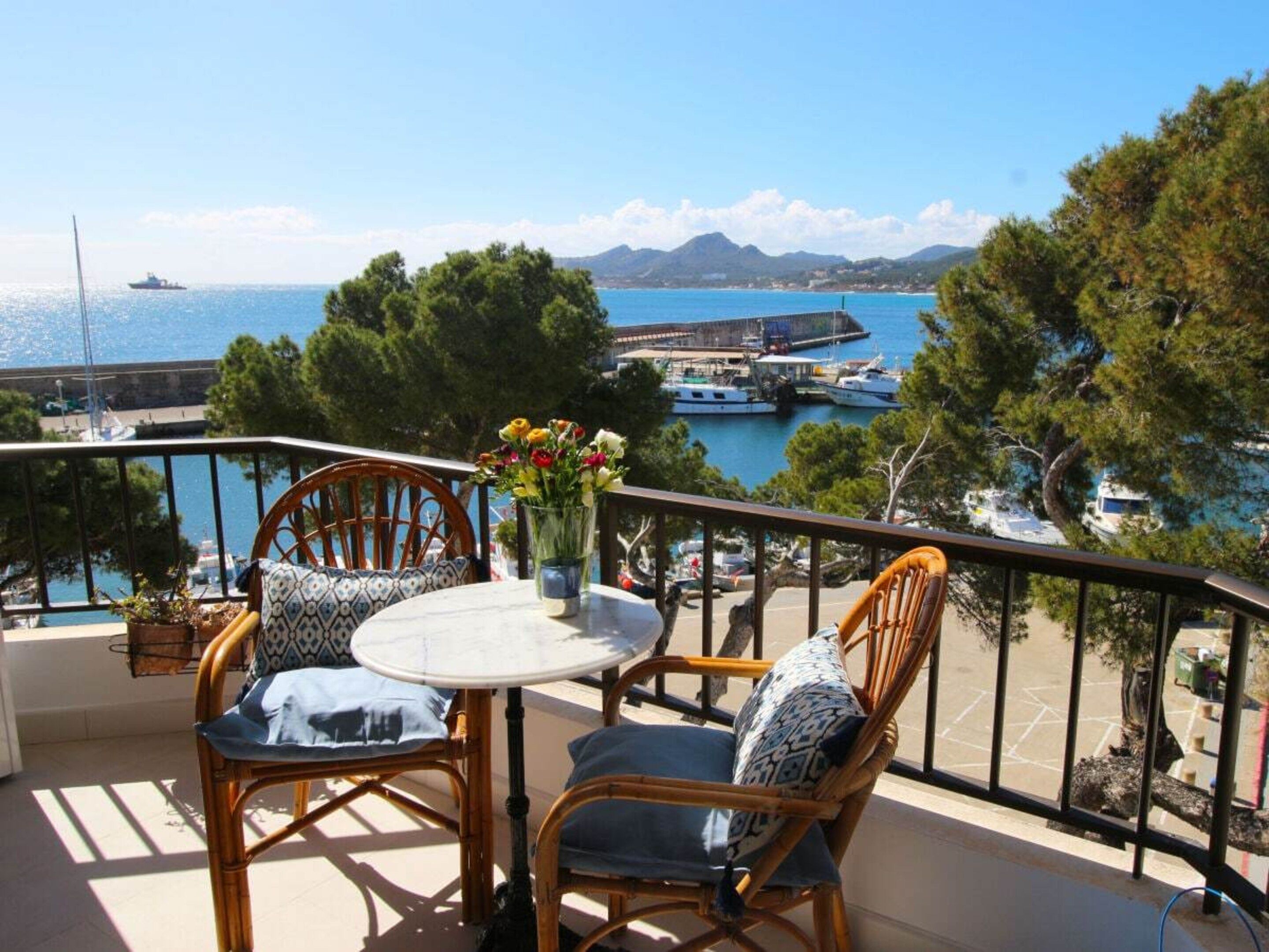 Un balcón con sillas de mimbre y una mesa, con vistas a un puerto deportivo con barcos y una montaña distante.