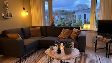 A modern living room with a sectional sofa, a round coffee table with decorative items, a flat-screen TV on a stand, and a view of the neighborhood through large windows.