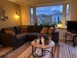Ein modernes Wohnzimmer mit Ecksofa, rundem Couchtisch mit Dekoration, Flachbildfernseher auf einem Standfuß und Blick auf die Nachbarschaft durch große Fenster.
