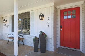 Una casa con puerta roja, contraventanas blancas y un porche con banco y plantas en macetas.