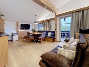 Ein modernes Wohnzimmer mit einer Couch, einem Essbereich mit Stühlen, einem an der Wand montierten Fernseher und großen Fenstern mit Vorhängen.
