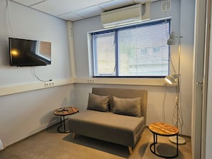 A small, modern room with a sofa, a TV mounted on the wall, a window with blinds, and a desk with a lamp. - We01 Het Kantoor (Westkapelle)