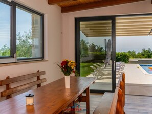 A modern outdoor seating area with a wooden table and benches, a glass door leading to a poolside, and a view of greenery. - Villa in Jursici With Swimming Pool (Svetvincenat)