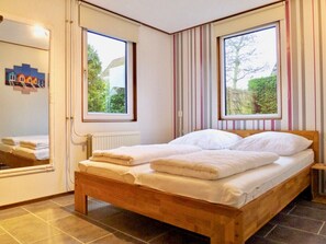 Ein Schlafzimmer mit einem hölzernen Bett, zwei Fenstern, einem Heizkörper und einem Spiegel.