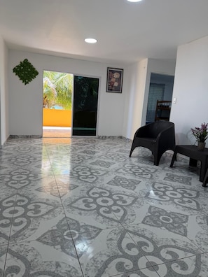 Una habitación amplia con suelo estampado, una silla negra, una mesita y una puerta que da a un espacio exterior con vistas a la vegetación.