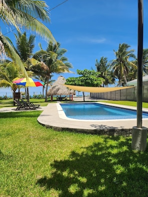 A swimming pool surrounded by grass and palm trees, with a thatched-roof structure and a colorful umbrella. - Beach Getaway in El Zapote - 3BR Home with AC (El Zapote)