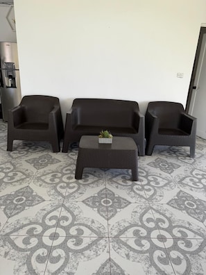 Un área de estar con tres sofás de color oscuro y una mesa de centro a juego, sobre baldosas con diseño.