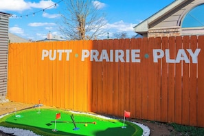 A wooden fence with 'PUTT PRAIRIE PLAY' painted on it, a miniature golf course with two flags, and a clear blue sky. - 15 Min to AT&T Stadium|FIFA 2026|Pool|Spa|Games (Grand Prairie)