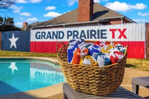 A wicker basket filled with colorful towels on a poolside deck with a pool in the background and a fence displaying 'GRAND PRAIRIE TX' with a star. - 15 Min to AT&T Stadium|FIFA 2026|Pool|Spa|Games (Grand Prairie)