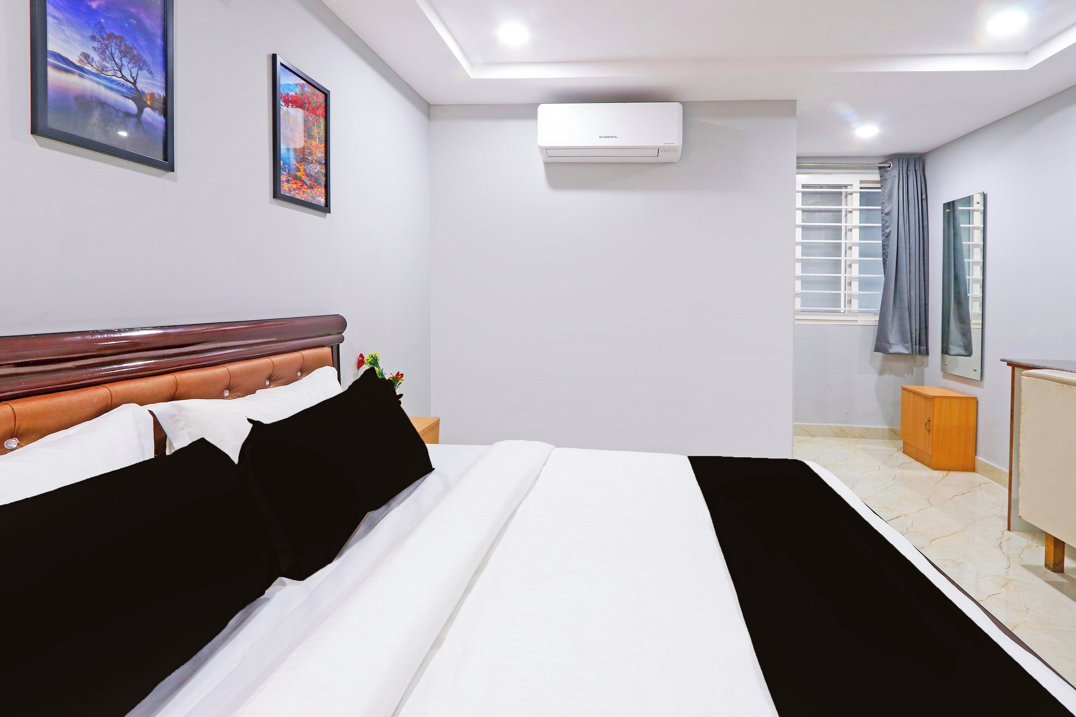 A hotel room with a bed, a nightstand, a window with curtains, an air conditioning unit, and artwork on the walls.