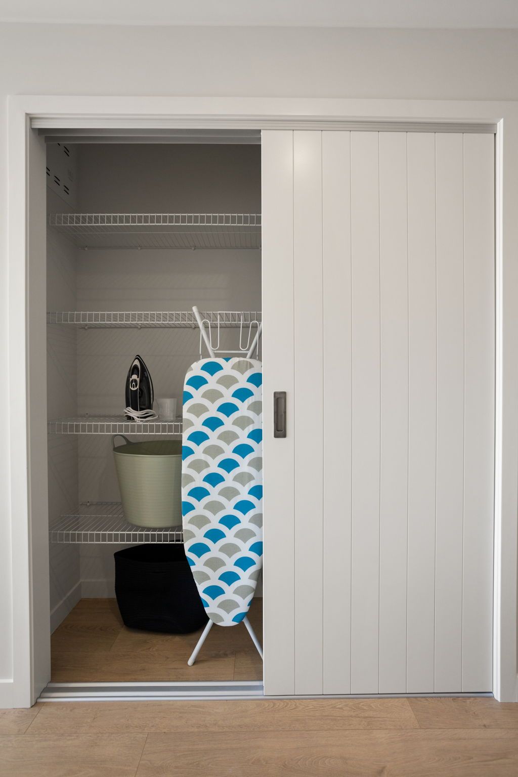 Een kamer met een witte garderobe, een betegelde muur, een plank met een waterkoker, een emmer en een gestreept strijkplank.