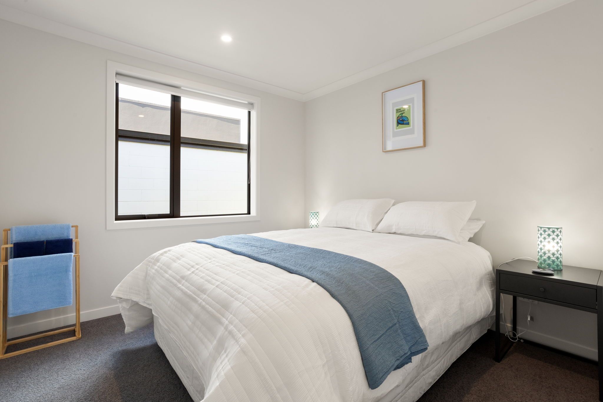 A bedroom with a bed, a bedside table, a nightstand, a window, and a towel rack.