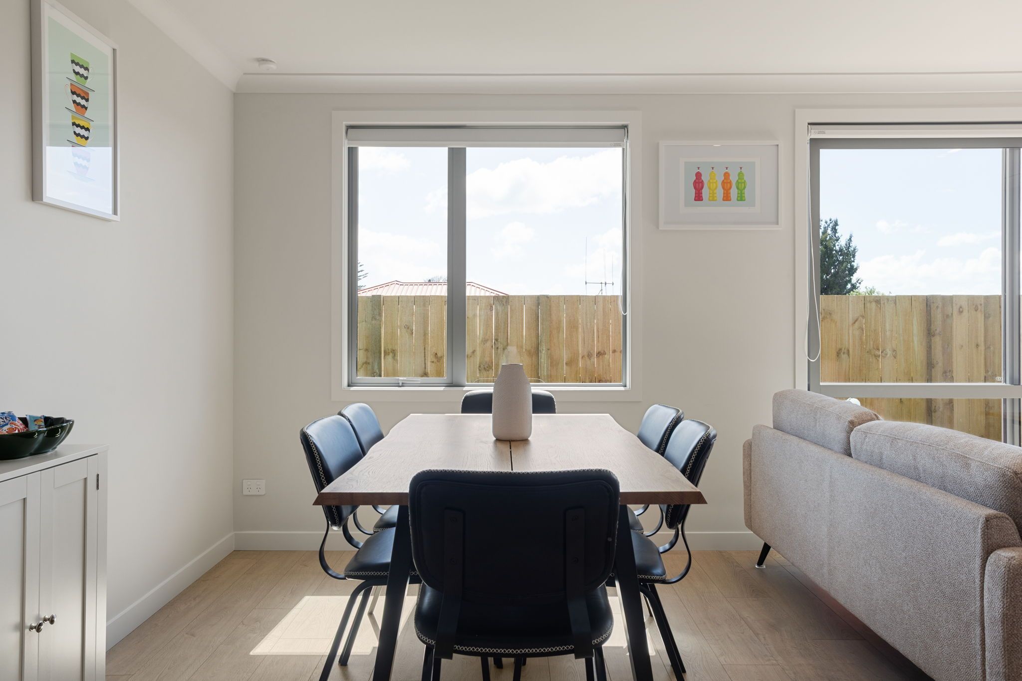 Un comedor moderno con una mesa, sillas, un sofá y ventanas amplias con vista a una cerca de madera.