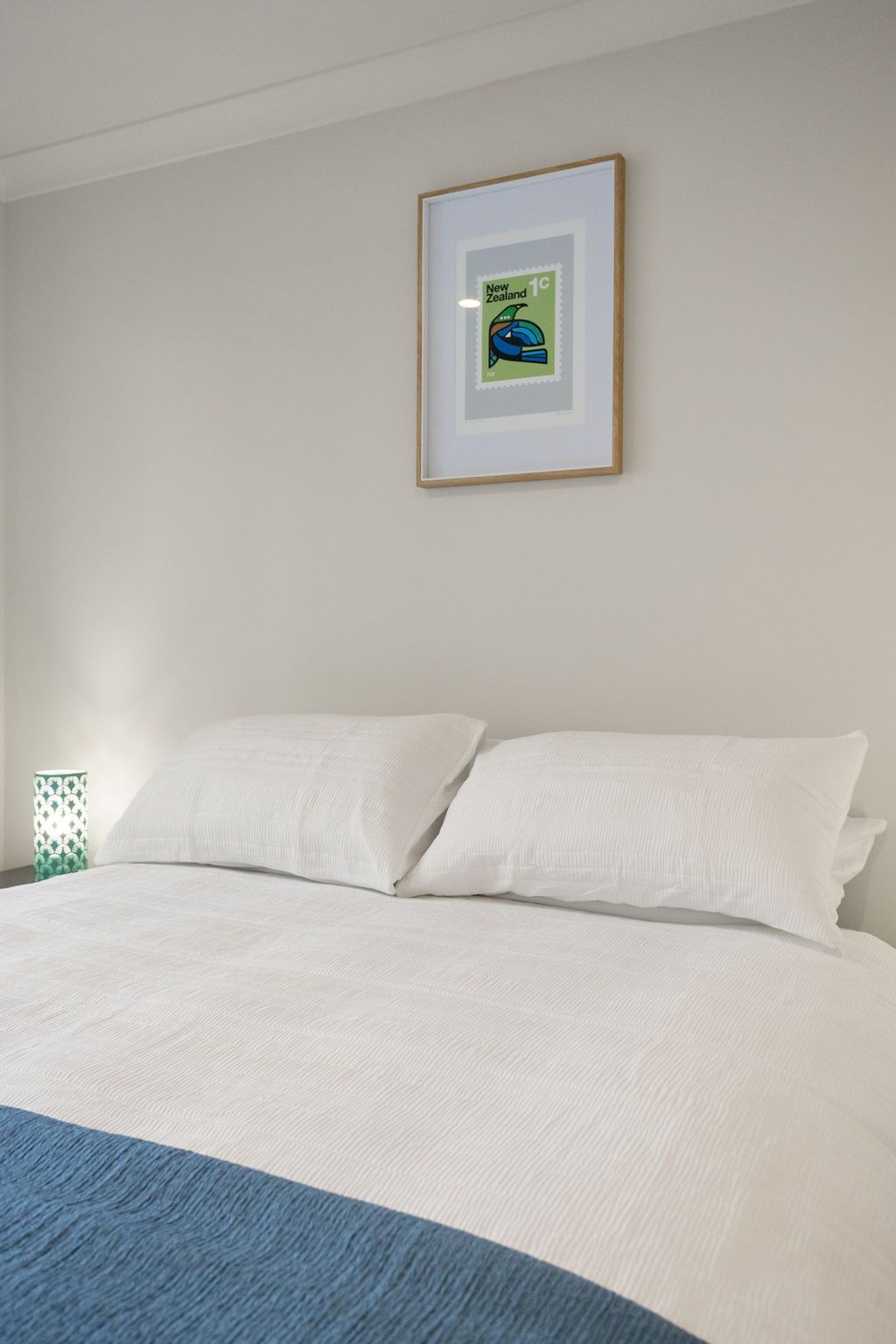 Een slaapkamer met een bed, witte kussens, een inlijsting foto aan de muur en een blauw detail bij het voeteneinde van het bed.