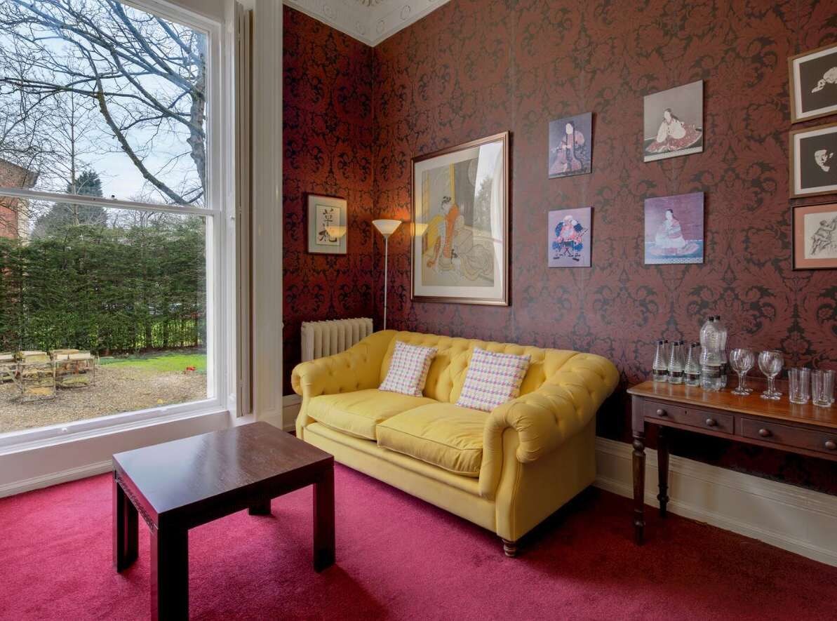 Une pièce avec un canapé jaune, une table en bois et un radiateur. Des tableaux encadrés ornent le mur, et une fenêtre offre une vue sur les arbres.