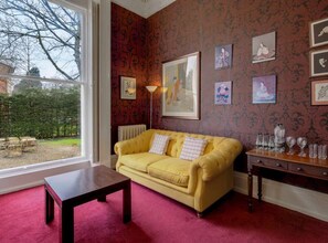 Une pièce avec un canapé jaune, une table en bois et un radiateur. Des tableaux encadrés ornent le mur, et une fenêtre offre une vue sur les arbres.