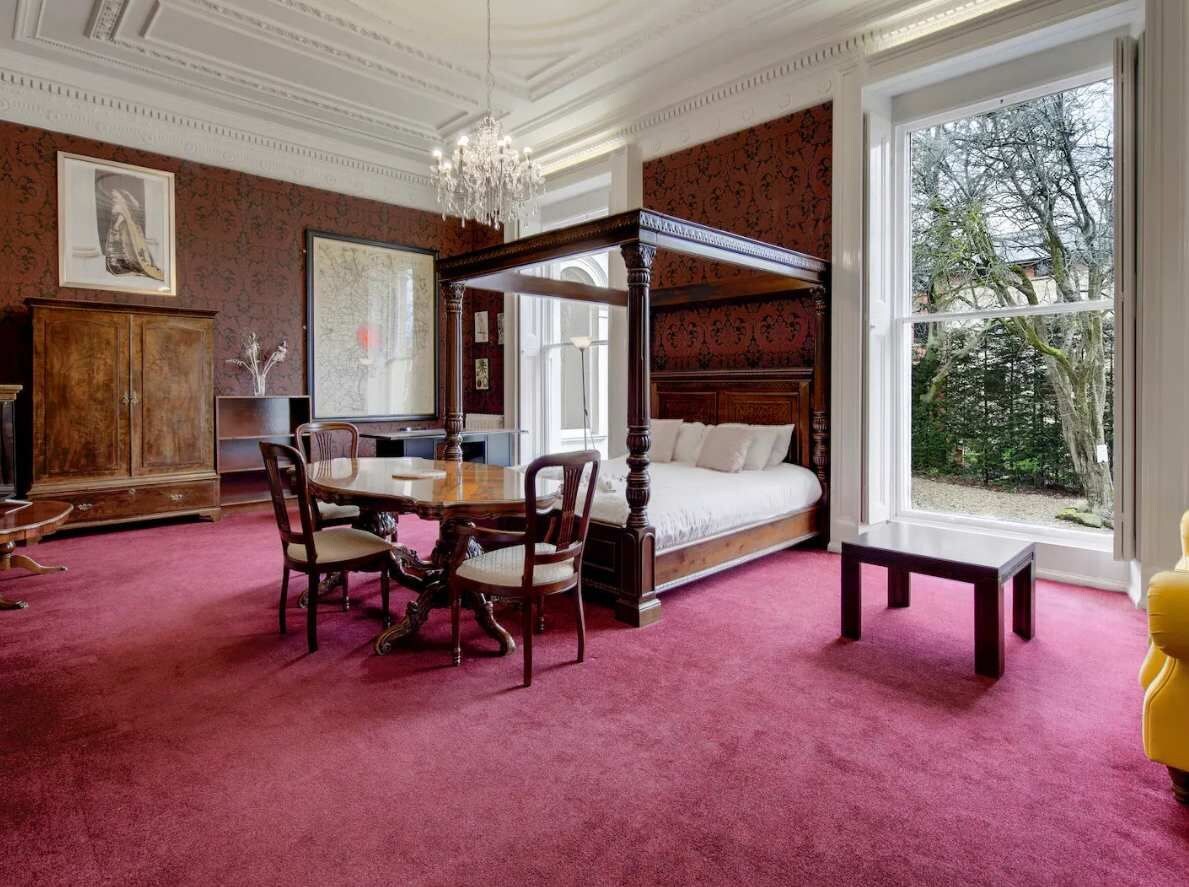 Een slaapkamer met een hemelbed, een eettafel met stoelen, een houten kast en een groot raam met uitzicht op bomen.