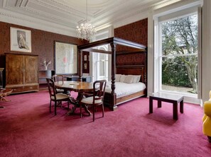 Une chambre à coucher avec un lit à baldaquin, une table à manger avec des chaises, un buffet en bois et une grande fenêtre donnant sur les arbres.