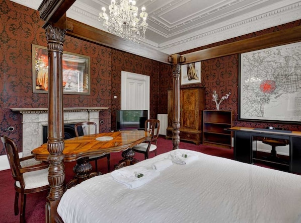 Une chambre d’hôtel avec un grand lit, une table à manger en bois avec des chaises, une cheminée, une carte accrochée au mur et un lustre.