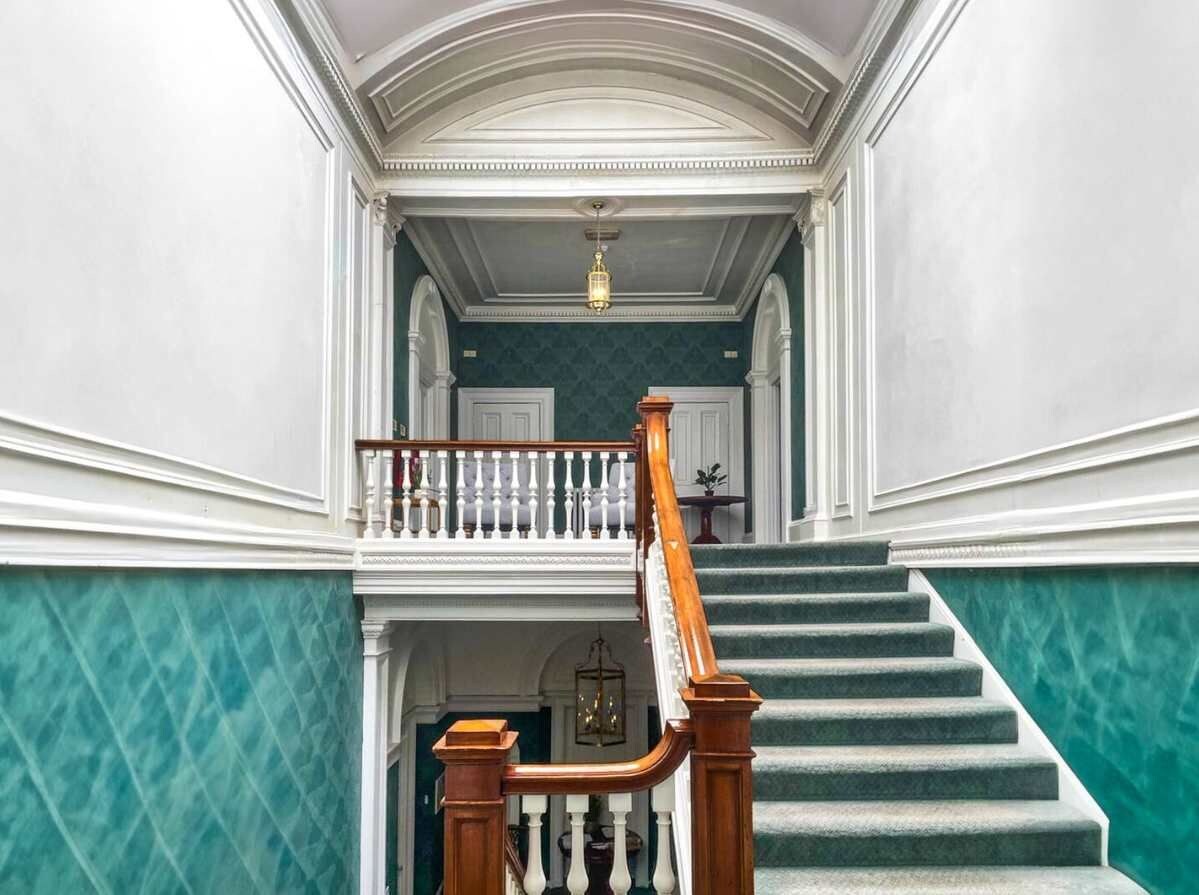 Un grand escalier en bois, doté d’une rampe et de balustres blanches, mène à un palier supérieur où est suspendu un lustre.