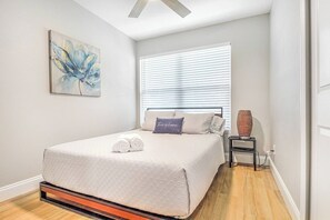 A bedroom with a bed, a window with blinds, a painting on the wall, and a bedside table with a lamp. - Lovely Modern POOL home. 13 mins to the Beach! (Melbourne)