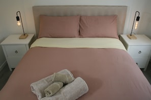 Ein Bett mit beiger und rosa Bettwäsche, zwei Nachttische mit Lampen und ein Paar Handtücher auf dem Bett.