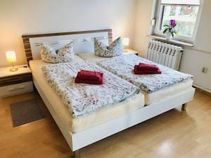 Ein Doppelbett mit floralem Bettbezug, zwei rote Handtücher, Nachttische mit Lampen, eine Heizung und ein Fenster mit einer Pflanze.