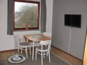 A room with a dining table and chairs, a radiator, a television mounted on the wall, and a window with curtains. - Holiday Apartment With 2 Bedrooms (Wangerooge)