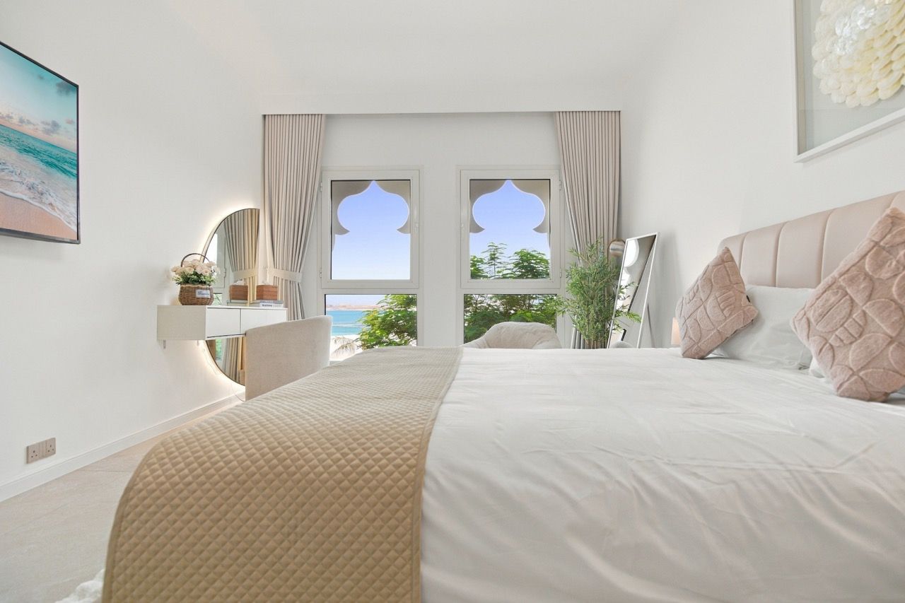 A bedroom with a large bed, a view of the sea through a window, and a bedside table with a lamp.
