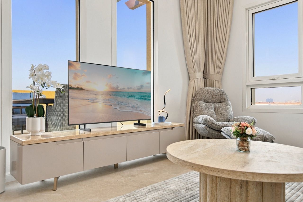 A modern living room with a flat-screen TV, a grey sofa, a round coffee table, and a view of the sea through large windows.