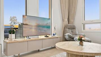 A modern living room with a flat-screen TV, a grey sofa, a round coffee table, and a view of the sea through large windows.