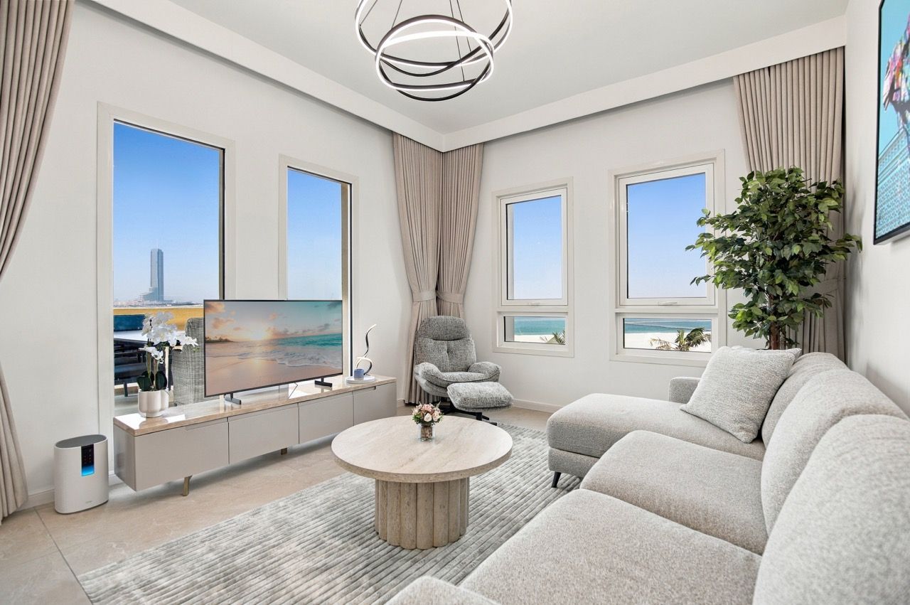 A modern living room with a large flat-screen TV, a grey sofa, a round coffee table, and a view of the cityscape through large windows.