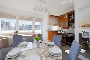 A modern kitchen with a city view, a dining area with a table set for four, and a large window with a view of buildings. - Warsaw | Solidarności Avenue 92 | Two-storey Apartment | Balcony (Warszawa)