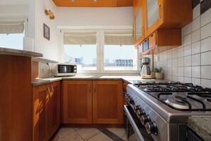 A modern kitchen with wooden cabinets, a gas stove, a microwave, and a window with a city view. - Warsaw | Solidarności Avenue 92 | Two-storey Apartment | Balcony (Warszawa)