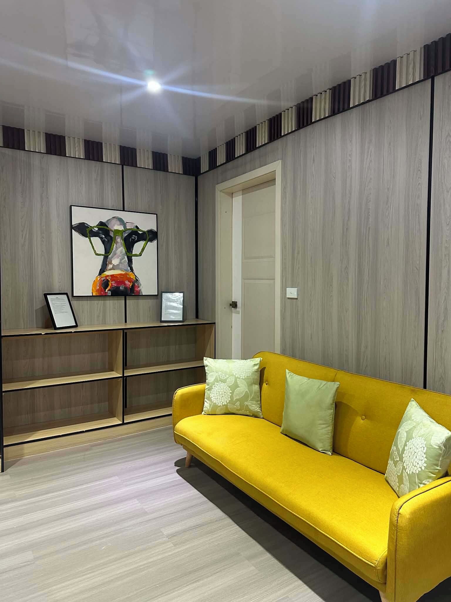 A modern living room with a yellow sofa, a framed artwork, and a wooden bookshelf.