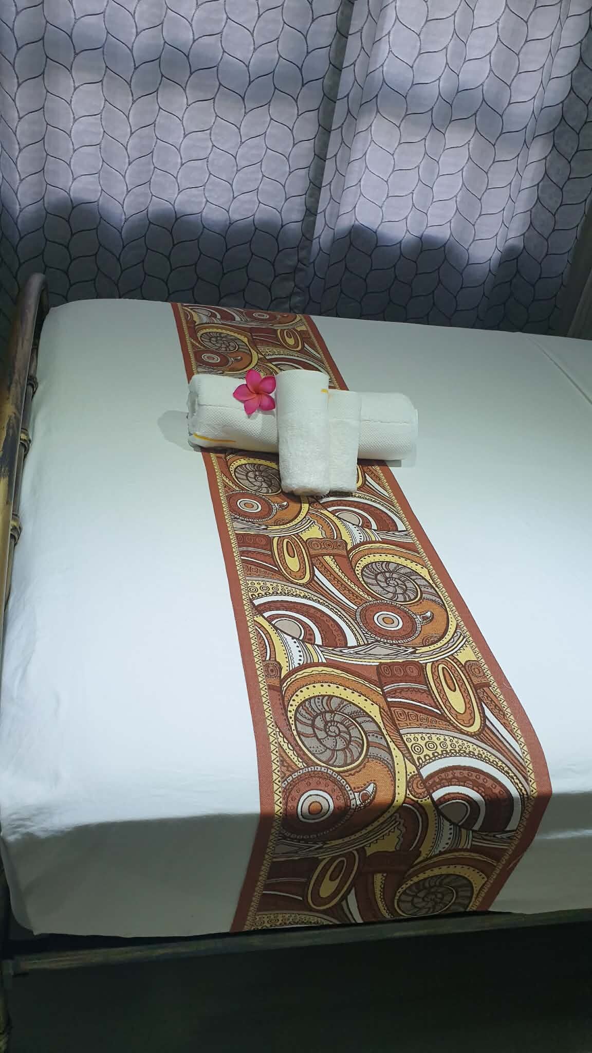 A bed with a patterned bedspread, a rolled towel, and a pink flower.