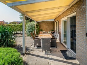An outdoor patio area with a covered table and chairs, a brick wall, and a glass door. - Holiday Home With 3 Bedrooms (Burgh-Haamstede)