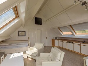 Ein Zimmer mit hohen Decken und schrägen Wänden, ausgestattet mit einem Bett, einer Sitzecke mit Stühlen, einer Küchenzeile und Oberlichtern.