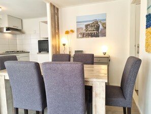 A dining area with a wooden table and grey upholstered chairs, a kitchen in the background, and a wall-mounted picture of a coastal scene. - Ok17 Holiday Home Oostkapelle (Oostkapelle)