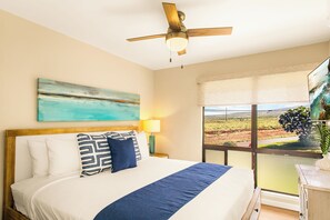 A bedroom with a bed, a ceiling fan, a painting, a lamp, and a window with a view. - Gorgeous Kaanapali 3BR with Dreamy Ocean Views (Lahaina)