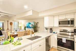 A modern kitchen with white cabinetry, stainless steel appliances, and a dining area with wicker furniture. - Gorgeous Kaanapali 3BR with Dreamy Ocean Views (Lahaina)