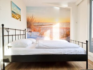 Ein Bett mit Metallgestell, weißer Bettwäsche und einem Wandbild, das einen Sonnenuntergang über einem Strand zeigt.