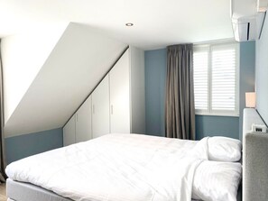 A bedroom with a sloped ceiling, a bed with white bedding, a window with curtains, and a wall-mounted air conditioning unit. - Ok10, Luxury, Quietly Located Holiday Home (Oostkapelle)